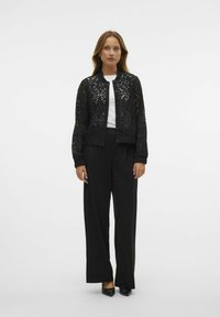 Veste bomber en dentelle noire avec des poignets et un ourlet côtelés, associée à un pantalon noir ample et une chemise blanche. Design et texture minimalistes.
