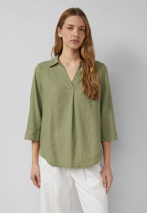 Vrouw met lang lichtbruin haar, gekleed in een olijfgroene V-hals blouse met driekwartmouwen en een witte broek, staand voor een grijze achtergrond.