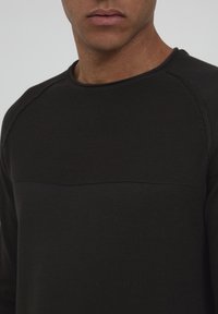 Chemise noire à manches longues avec un motif texturé et un panneau lisse sur la poitrine, dotée d'un col rond et de détails de couture contrastants.