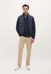 Gilet imbottito blu navy su una maglia a maniche lunghe blu navy, abbinato a pantaloni beige chiaro e sneakers bianche, in piedi contro uno sfondo semplice.