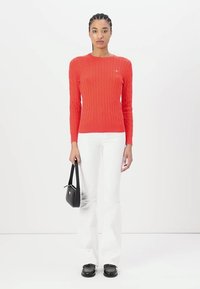 Suéter rojo de punto trenzado con cuello redondo, mangas largas, combinado con pantalones de mezclilla blancos acampanados y un bolso negro. Textura suave y diseño ajustado.