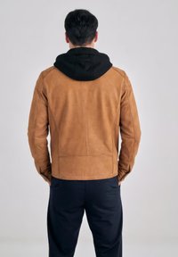 Uomo in piedi con le mani nelle tasche, indossa una giacca di pelle color cuoio sopra una maglia nera con cappuccio e pantaloni scuri, di spalle su uno sfondo semplice.