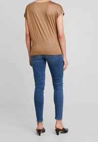 Hnědý top s krátkým rukávem vyrobený z hladkého materiálu, doplněný modrými skinny džíny. Černé boty na vysokém podpatku s mírným podpatkem zdůrazňují celý outfit.