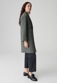 Grau melierter Wollmix-Mantel mit strukturiertem Design, zwei Knöpfen und großen Seitentaschen, kombiniert mit dunklen Jeanshosen und schwarzen Slippers.