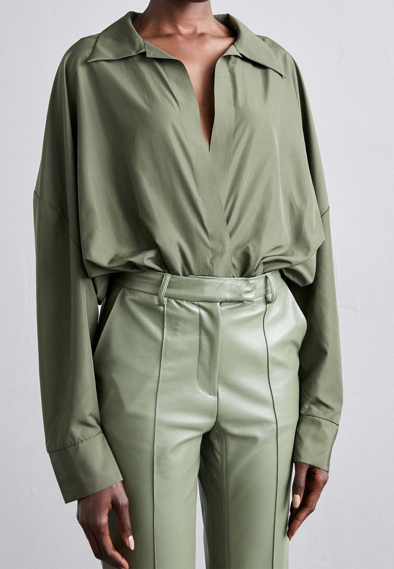 Chemise oversize vert olive avec col, dotée d'un décolleté en V profond et d'une taille froncée. Associée à un pantalon en cuir vert brillant taille haute.