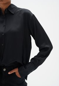 Schwarzes Langarmhemd mit Button-Down-Kragen, aus glattem Stoff gefertigt. Mit vorderen Knöpfen und leicht lockerer Passform an den Handgelenken.