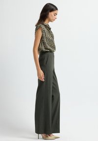 Femme portant un haut sans manches à motifs, un pantalon large taille haute sombre et des escarpins beiges à bout pointu, se tenant contre un fond blanc.