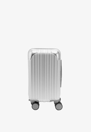 Srebrna walizka twarda z pionowymi rowkami, wzmocnionymi narożnikami i czterema obrotowymi kołami, stojąca pionowo na białym tle.