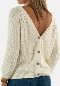 Pull en maille crème avec un dos en V profond, comportant quatre boutons décoratifs. Le motif texturé et les poignets côtelés ajoutent de l'intérêt visuel.