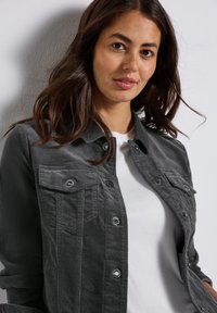 Šedá džínsová bunda s předními kapsami, stříbrnými cvoky a měkkou texturou. Nosená přes bílý tričko, s jemným leskem.