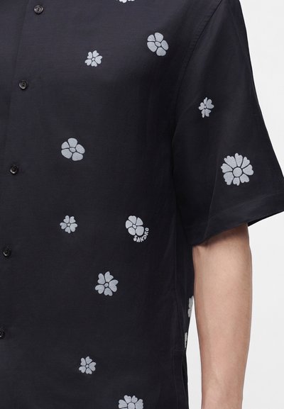Chemise noire à manches courtes avec de petits motifs floraux blancs dispersés sur le tissu, portée par une personne avec l’avant-bras visible.