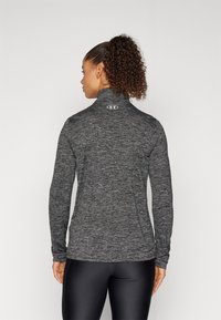 Frau mit lockigem Pferdeschwanz, die ein eng anliegendes, dunkelgraues langärmliges Sportoberteil und schwarze Leggings trägt, steht mit dem Rücken zu einem schlichten Hintergrund.