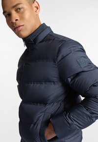 Marine blauwe pufferjack met hoge kraag, rits aan de voorkant en geribde manchetten. Voorzien van een logopatch op de linker mouw en een gladde, glanzende textuur.
