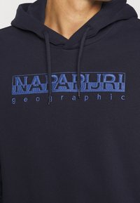 Felpa navy con tasca a marsupio, caratterizzata dal logo ricamato "NAPAPIJRI" in blu e la scritta "geographic" in testo più piccolo sottostante. Tessuto morbido.