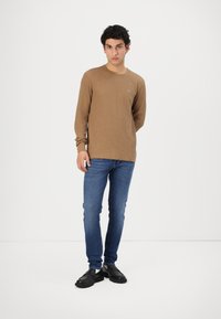 Barna hosszú ujjú ing texturált mintával, kék slim-fit farmernadrággal és fekete mokaszin cipővel párosítva. Egyszerű logó a mellkason.