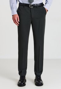 Mann in dunkelgrauen Anzughosen, schwarzem Gürtel, schwarzen dressierten Schuhen, hellblauem Hemd und gemustertem Krawatte, steht mit den Händen in den Taschen.