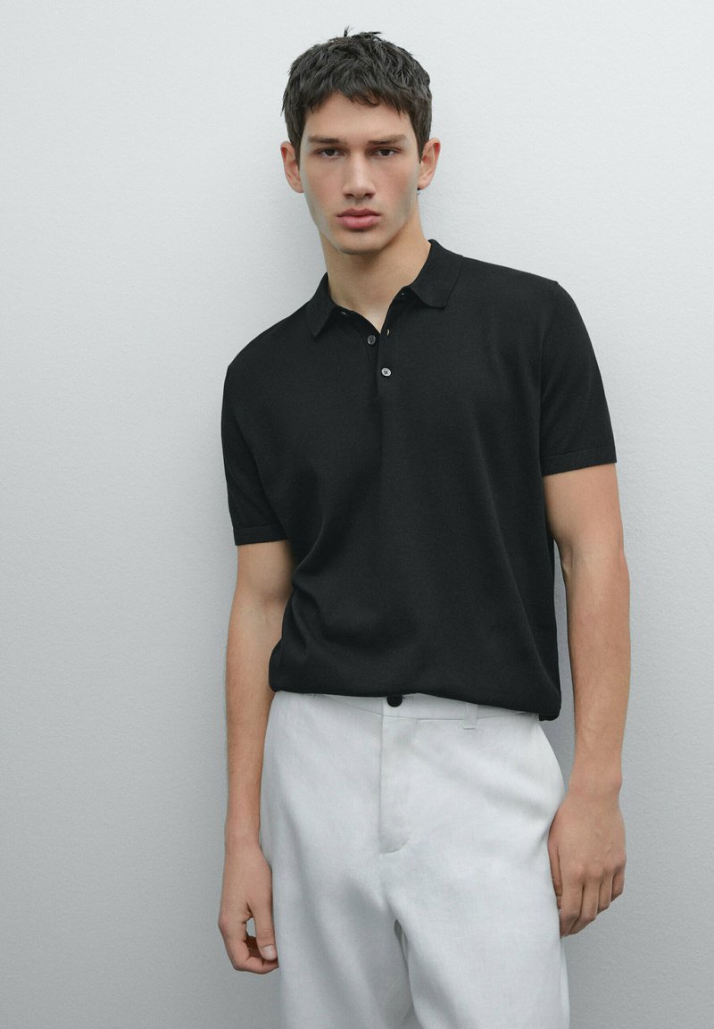 Polo shirt nero a maniche corte con pattina a tre bottoni, texture liscia e design aderente, abbinato a pantaloni chiari.