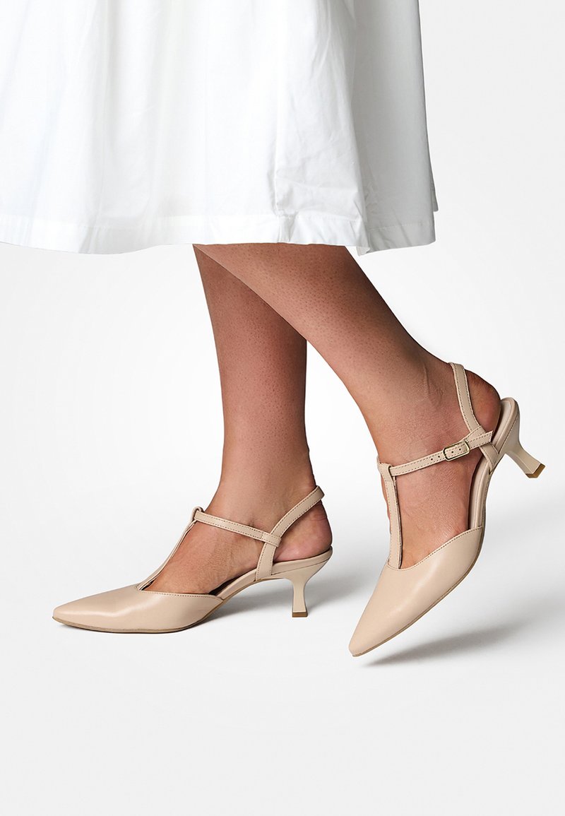 Escarpins beige à bout pointu et brides arrière avec un design en T, petits talons carrés et finition en cuir lisse. Accentué par des sangles de cheville réglables.