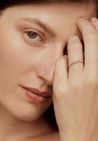 Primer plano de una mujer con piel clara mostrando un anillo de oro con pequeños diamantes en su dedo medio cerca de su cara.