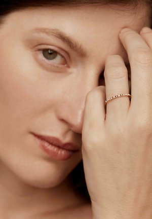 Primer plano de una mujer con piel clara mostrando un anillo de oro con pequeños diamantes en su dedo medio cerca de su cara.