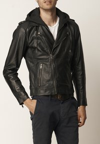 Chaqueta de motocicleta de cuero negro con costuras decorativas, bolsillos con cremallera y capucha gris removible. Corte ajustado y textura suave.