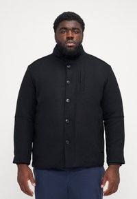 Black, button-up coat with high collar; made of a textured fabric. Features side pockets and a subtle seam detail on shoulders.
