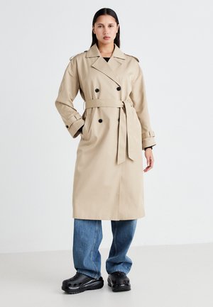 Beige trench coat with double-breasted front, belted waist, and shoulder epaulettes; features wide sleeves and pockets; paired with blue jeans.