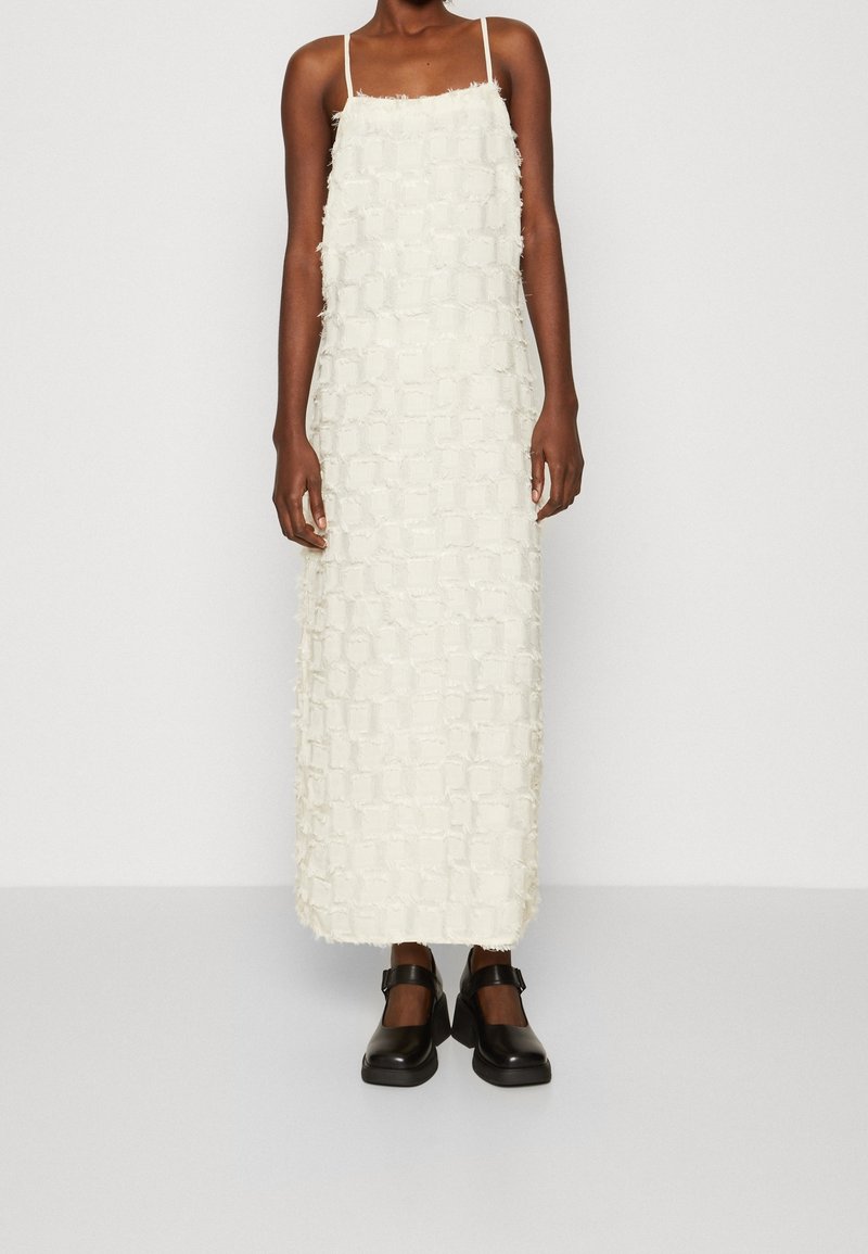 Kobieta ubrana w białą, teksturowaną, bezrękawnikową sukienkę midi z cienkimi ramiączkami oraz czarne, grube buty Mary Jane na tle jednolitego tła.