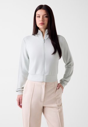 Femme aux longs cheveux noirs portant une veste zippée gris clair et un pantalon taille haute beige, debout avec une main dans la poche.