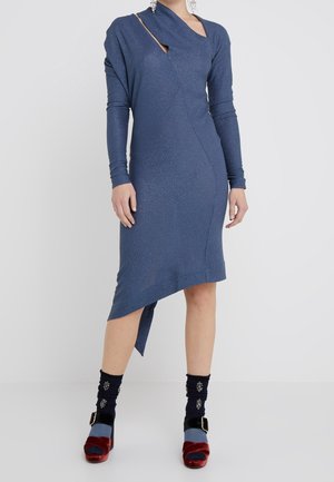 Mujer vestida con un vestido azul asimétrico de manga larga con un corte cerca del hombro, combinado con calcetines negros y tacones de plataforma de terciopelo rojo.