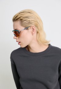 Occhiali da sole con montatura angolare nei toni del blu e del verde, dotati di lenti arancioni tintate. Indossati con una felpa grigia a girocollo.