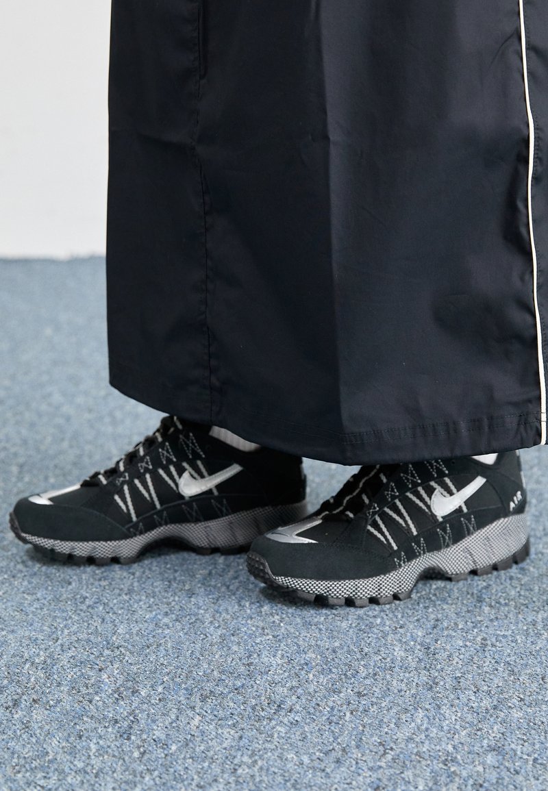 Des chaussures de sport noires avec un dessus texturé, des accents argentés et une semelle extérieure à motifs, associées à un long vêtement noir. Présentées sur un tapis bleu.