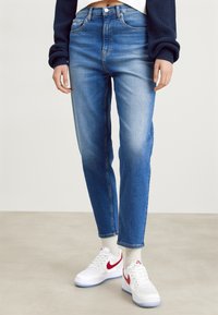 Jeans de denim azul con cintura ajustada y piernas rectas, con un ligero desgaste. Combinados con zapatillas blancas con acentos rojos.