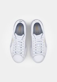 Baskets blanches en cuir lisse avec un design bas, comprenant une fermeture à lacets et des accents dorés sur les côtés et la languette.