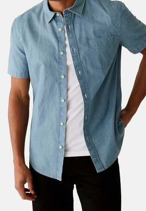 Mann trägt ein hellblaues, kurzärmeliges Jeanshemd über einem weißen T-Shirt und schwarzen Hosen, die Hand ruht auf der Hüfte.