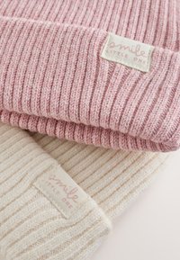 Kaksi taiteltua ribbineulottua pipoa vaaleanpunaisena ja kermanvärisenä, joissa kangasmerkit, joissa lukee "smile little one", yksivärisellä taustalla.