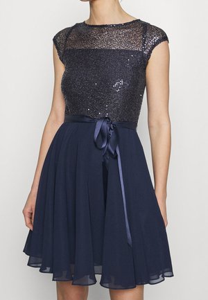 Vrouw draagt een donkerblauwe jurk zonder mouwen met een lijfje versierd met pailletten, doorschijnend bovenstuk op de borst en een satijnen lint gestrikt in de taille.