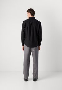 Hombre de espaldas vistiendo una camiseta de manga larga negra, pantalones grises y zapatos negros, contra un fondo claro y liso.
