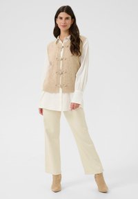 Ženska stoji v bež pletenem jopiču z mašnicami, oblečena v belo dolgo srajco, kreme barve hlače in bež gležnjarje, proti enotni beli ozadju.