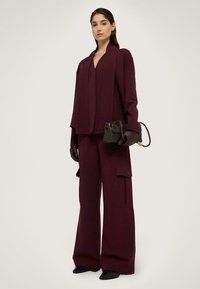 Bordeaux maatwerk blazer en wijde broek in een textuurstof. Accessoires omvatten een kleine zwarte handtas en bijpassende handschoenen.