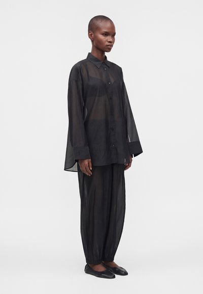 Femme aux cheveux courts debout, portant une chemise noire ample et transparente à boutons et un pantalon large assorti avec des ballerines noires sur un fond uni.