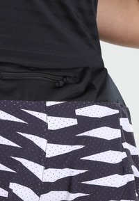 Sportig tanktop i svart med horisontella ränder, har en svart dragkedjeficka; den nedre delen har en mönstrad design i vitt och grått.