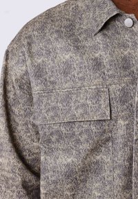 Veste chemise à motifs gris et beige, dotée d'un col pointu, d'une fermeture à boutons-pression et de deux poches poitrine. Fabriquée en tissu texturé.