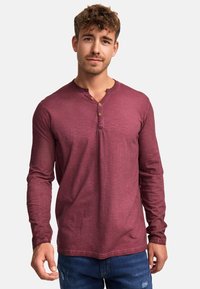Langärmliges, burgunderfarbenes Henley-Shirt mit rundem Halsausschnitt, Knopfleiste mit vier Knöpfen und strukturierter Stoffoberfläche, kombiniert mit blauen Jeans.