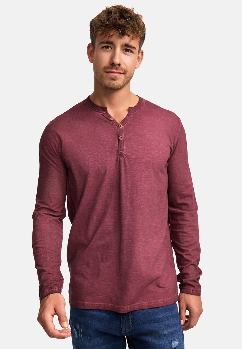 Langärmliges, burgunderfarbenes Henley-Shirt mit rundem Halsausschnitt, Knopfleiste mit vier Knöpfen und strukturierter Stoffoberfläche, kombiniert mit blauen Jeans.