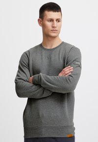Sudadera gris hecha de tela suave, con cuello redondo, mangas largas y un parche de logo en la manga izquierda. Puños y dobladillo acanalados.