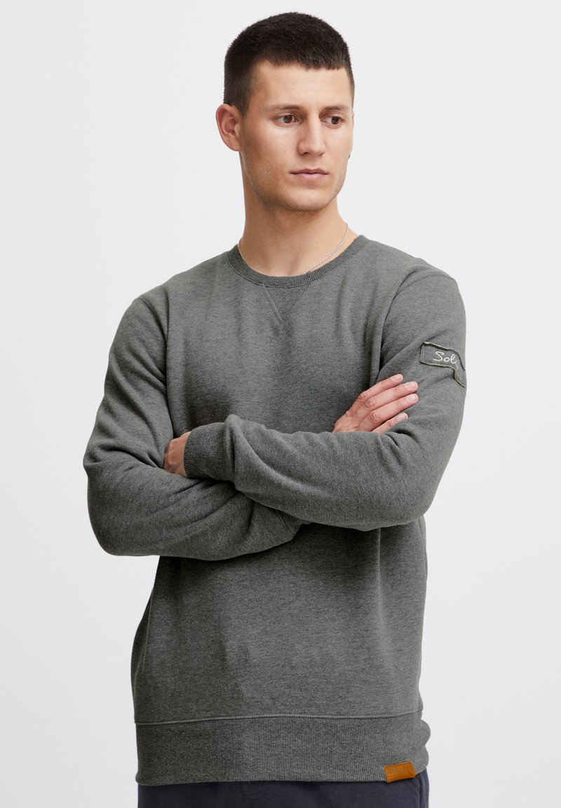 Sudadera gris hecha de tela suave, con cuello redondo, mangas largas y un parche de logo en la manga izquierda. Puños y dobladillo acanalados.