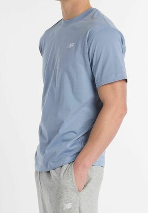 Mann trägt ein hellblaues kurzärmeliges Hemd und hellgraue Jogginghosen, steht mit einer Hand in der Tasche vor weißem Hintergrund.