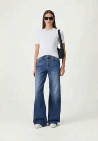 Woman wearing black sunglasses, white short-sleeve shirt, wide-leg blue jeans, white shoes, and a black shoulder bag on a plain background.