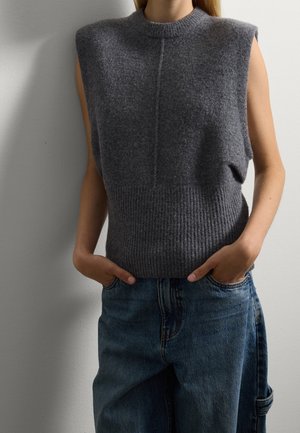 Person, die eine ärmellose dunkelgraue Strickweste und blaue Jeans trägt, mit den Händen in den Taschen, stehend vor einer schlichten hellen Wand.